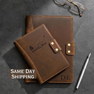 May include: A dark brown leather notebook and a smaller matching leather passport holder with gold-tone snap closures. The passport holder features an engraved airplane icon, a heart, and the text DANIEL|EVA. The notebook is engraved with DE.