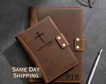 May include: Two brown leather journals, one small and one large, with a cross and the text "Let there be light." Genesis 1:3. The smaller journal has a snap closure. A silver pen and eyeglasses are also visible.
