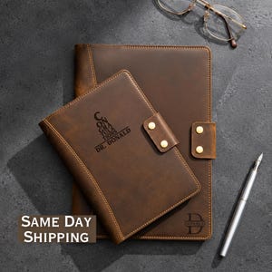 May include: Two brown leather journals with gold-tone snap closures. The smaller journal features the engraved text "CONGRATULATIONS DR. DONALD" and the larger journal features the engraved letter "D" with the name "DONALD" below it.