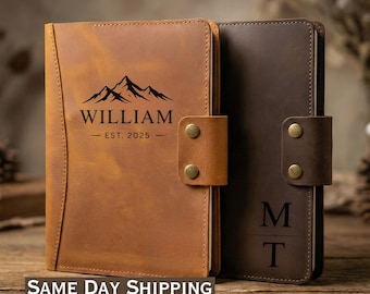 May include: Two leather journals, one tan and one dark brown, are displayed on a wooden surface. The tan journal features a mountain graphic with the name "WILLIAM" and "EST. 2025". Both journals have a snap closure.