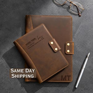May include: A brown leather notebook and a matching leather portfolio featuring gold-tone snap closures. The notebook is engraved with the text MARK TWIN boss YOU ARE THE EVERYONE WISHES THEY HAD, and the portfolio features the initials MT.