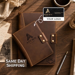 May include: Two brown leather notebook covers with gold-tone metal snap closures. The smaller cover features the embossed logo ADEWN REALTY, while the larger cover displays the initials DK above an antler graphic. A silver fountain pen lies nearby.