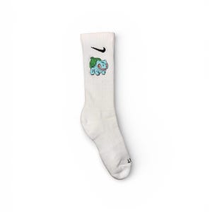 Op de afbeelding: Witte sportsok met een zwarte Nike swoosh en een Bulbasaur-afbeelding in blauw, groen en rood. De sok is een crew-lengte en heeft een geribbelde textuur.