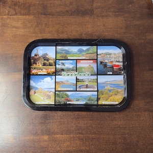 Puede incluir: Bandeja rectangular negra con varias imágenes de paisajes y monumentos de Escocia. La bandeja incluye imágenes de castillos, montañas y barcos, con la palabra "Scotland" en el centro.