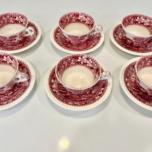 Może przedstawiać: Zestaw sześciu czerwono-białych ceramicznych filiżanek do herbaty z pasującymi spodkami, każda z nich posiada szczegółowy wzór kwiatowy i krajobrazowy, faliste brzegi spodków oraz ozdobne ucho przy każdej filiżance, ułożone w dwóch rzędach po trzy.
