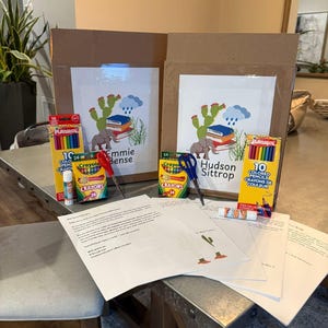 Puede incluir: Dos cajas de cartón con obras de arte personalizadas, crayones, lápices de colores, barras de pegamento y tijeras. La obra de arte presenta un cactus, un elefante, libros y los nombres "Emmie Bense" y "Hudson Sittrop".