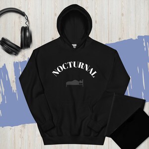 May include: Black hooded sweatshirt with the word "NOCTURNAL" in white arched text above a bed graphic. The sweatshirt has a front pocket and a drawstring hood. A pair of black headphones is in the background.