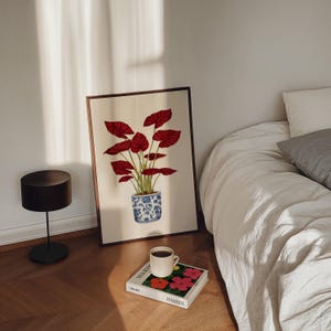 Puede incluir: Una impresión artística enmarcada con una planta de hojas rojas en una maceta con estampado azul y blanco. La obra de arte está apoyada contra una pared junto a una cama con ropa de cama blanca. Un libro y una taza de café están en el suelo.