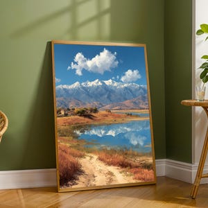 May include: A framed art print depicting a mountain range with snow-covered peaks reflected in a lake. The artwork shows a blue sky with white clouds, and a dirt track leads to the water.