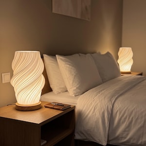 Puede incluir: Dos lámparas de mesa blancas con una textura cilíndrica, espiral y acanalada, con una base de madera clara. Las lámparas emiten un brillo cálido y suave, iluminando las mesitas de noche a ambos lados de una cama con ropa de cama blanca.