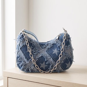 May include: A blue denim shoulder bag featuring a quilted diamond pattern with star motifs. The bag has frayed edges, a silver-toned metal chain strap, and a top zipper closure. The construction includes light blue piping along the seams.