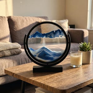 May include: A circular black frame containing a dynamic sand art display with layers of blue and white sand forming mountain-like peaks and valleys. The frame is mounted on a rectangular black base, creating a rotating visual landscape.