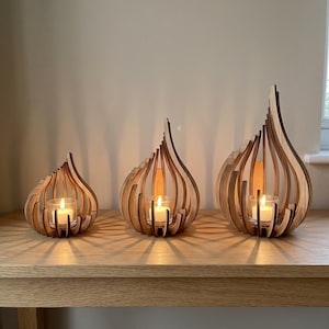May include: Three wooden candle holders of varying heights, each featuring a curved, flame-like slatted design. Each holder contains a clear glass votive cup with a lit white tea light candle inside, casting light patterns onto the surface below.