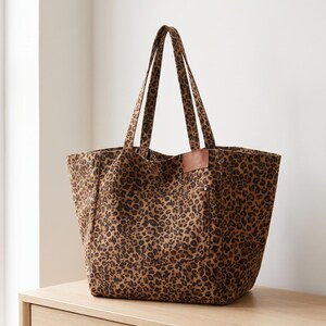 May include: A tote bag featuring a brown and black leopard print pattern. The bag has two shoulder straps, a rectangular brown leather-like patch on the front, and visible metal rivets at the top corners of the front pocket area.