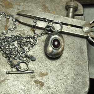 May include: A silver necklace with a toggle clasp and a pendant. The pendant is oval-shaped with a red center and a small white accent. The necklace and pendant are on a textured metal surface.
