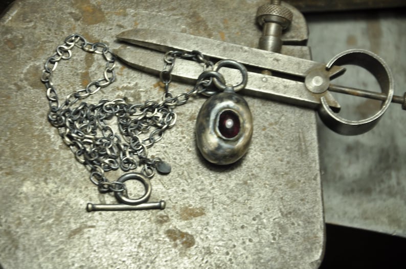 May include: A silver necklace with a toggle clasp and a pendant. The pendant is oval-shaped with a red center and a small white accent. The necklace and pendant are on a textured metal surface.