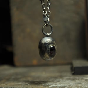 May include: A silver-toned pendant necklace featuring a textured, oval-shaped charm with a dark, eye-like stone. The pendant hangs from a delicate chain, creating a unique and artistic accessory.
