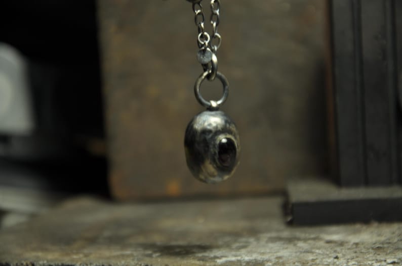May include: A silver-toned pendant necklace featuring a textured, oval-shaped charm with a dark, eye-like stone. The pendant hangs from a delicate chain, creating a unique and artistic accessory.