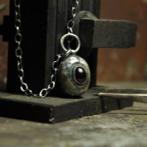 May include: A silver pendant necklace with an oval-shaped pendant featuring a dark red stone. The pendant hangs from a delicate chain. The necklace is displayed on a dark, textured surface, with a pair of tweezers in the foreground.