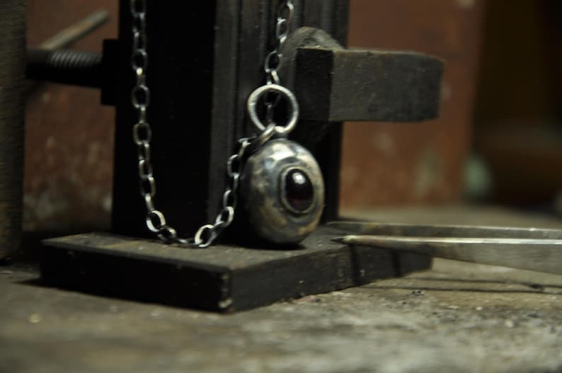 May include: A silver pendant necklace with an oval-shaped pendant featuring a dark red stone. The pendant hangs from a delicate chain. The necklace is displayed on a dark, textured surface, with a pair of tweezers in the foreground.