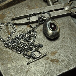 May include: A silver-toned necklace with a toggle clasp and a pendant. The pendant is oval-shaped with a red-toned center and a small, white pearl. The chain is made of small, textured links. The necklace is displayed on a textured, gray metal surface.