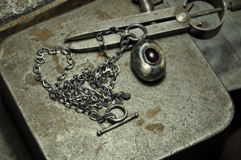 May include: A silver-toned necklace with a toggle clasp and a pendant. The pendant is oval-shaped with a red-toned center and a small, white pearl. The chain is made of small, textured links. The necklace is displayed on a textured, gray metal surface.