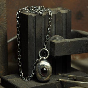 May include: A silver pendant necklace with a dark red gemstone set in a rounded, textured silver pendant. The pendant hangs from a silver chain. The necklace rests on a dark metal object, with a pair of metal tools in the foreground.