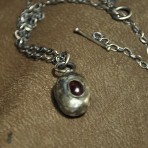 May include: A silver-toned necklace with a pendant featuring a red gemstone. The pendant is oval-shaped and set in a textured metal casing. The chain is a linked design with a toggle clasp.