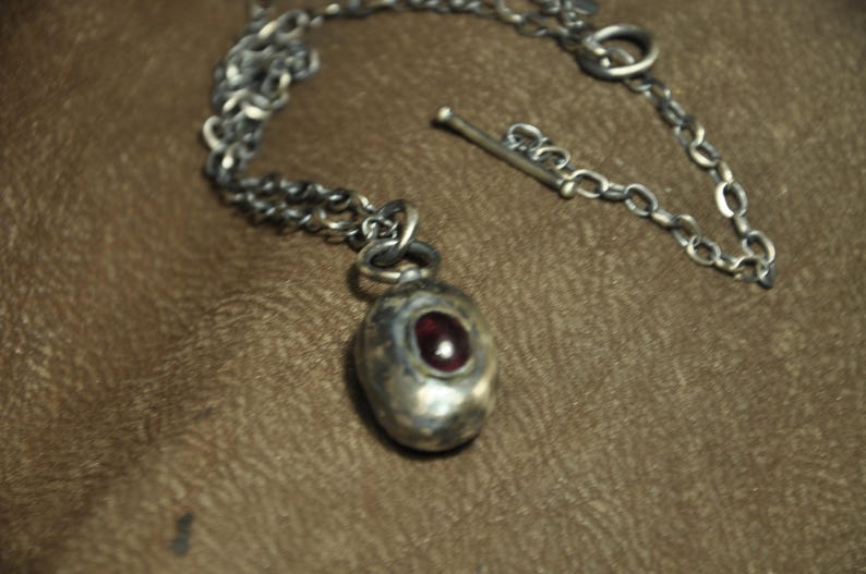 May include: A silver-toned necklace with a pendant featuring a red gemstone. The pendant is oval-shaped and set in a textured metal casing. The chain is a linked design with a toggle clasp.