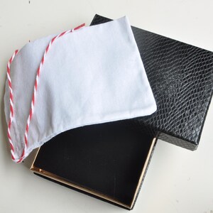 May include: A white fabric item with red and white trim, possibly a hat or accessory, rests atop an open black box with a gold-colored trim. The box is next to a black box with a textured, reptile-like pattern.