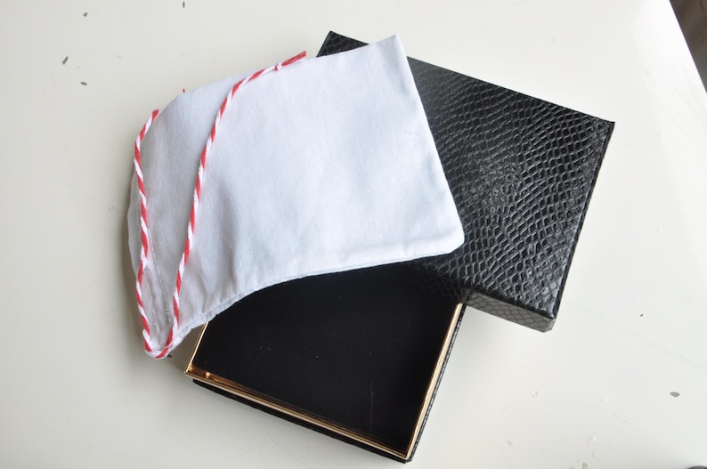 May include: A white fabric item with red and white trim, possibly a hat or accessory, rests atop an open black box with a gold-colored trim. The box is next to a black box with a textured, reptile-like pattern.