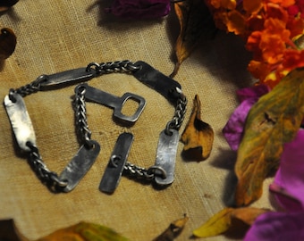 Könnte beinhalten: Ein silberfarbenes Metallarmband mit rechteckigen Gliedern und einem Flaschenöffner-Anhänger. Das Armband liegt auf einer strukturierten, beigen Oberfläche, umgeben von bunten Blumen und Herbstblättern.