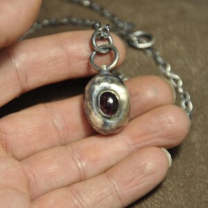 May include: A silver pendant necklace featuring a textured, oval-shaped pendant with a dark red gemstone. The pendant hangs from a silver chain. The necklace is held in a hand.