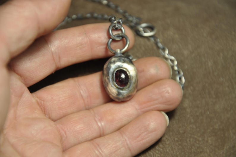 May include: A silver pendant necklace featuring a textured, oval-shaped pendant with a dark red gemstone. The pendant hangs from a silver chain. The necklace is held in a hand.