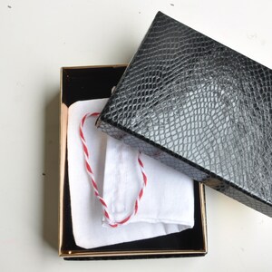 May include: An open black box with a gold trim, containing a white pouch with a red and white string. The box lid is black with a textured, reptile-like pattern. The box is on a white surface.