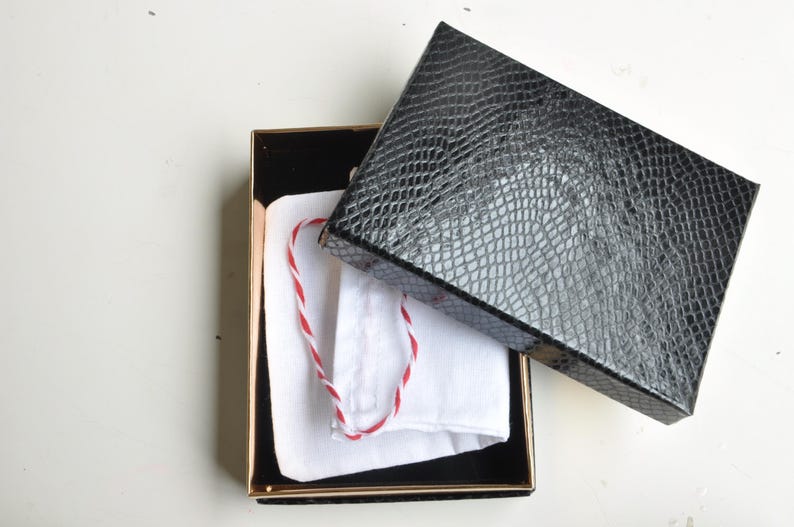 May include: An open black box with a gold trim, containing a white pouch with a red and white string. The box lid is black with a textured, reptile-like pattern. The box is on a white surface.