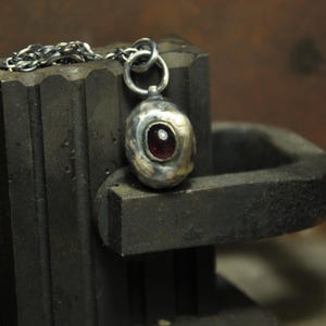 May include: A handcrafted silver pendant with a dark red gemstone, suspended from a delicate chain. The pendant is oval-shaped and set against a dark, textured background, showcasing the jewelry's unique design.