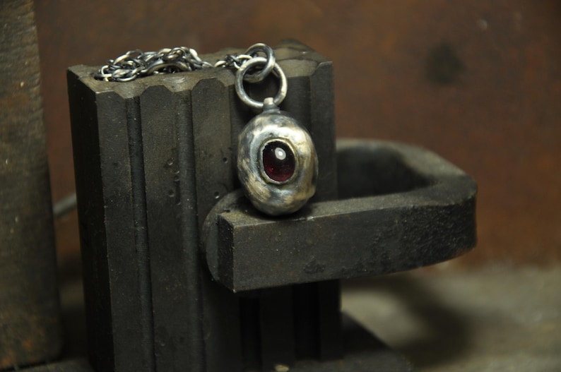 May include: A handcrafted silver pendant with a dark red gemstone, suspended from a delicate chain. The pendant is oval-shaped and set against a dark, textured background, showcasing the jewelry's unique design.