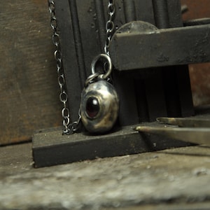 May include: A silver pendant necklace with a dark chain. The pendant is oval-shaped with a dark red stone in the center. The necklace is displayed on a dark, textured surface, with tools in the background.