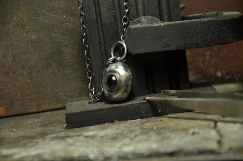 May include: A silver pendant necklace with a dark chain. The pendant is oval-shaped with a dark red stone in the center. The necklace is displayed on a dark, textured surface, with tools in the background.