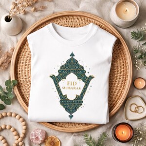 Peut inclure: T-shirt blanc avec un motif orné de couleur teal et or, portant l'inscription "Eid Mubarak". Le motif est centré sur le devant. Le t-shirt est plié sur un plateau tressé, entouré de bougies et d'éléments décoratifs.