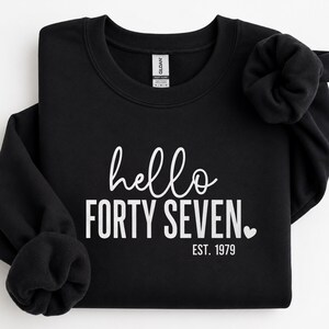 May include: A black crewneck sweatshirt featuring white screen-printed text that reads hello FORTY SEVEN. EST. 1979 with a small heart icon. The garment has a ribbed collar and long sleeves with gathered cuffs, displaying a Gildan brand label.