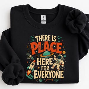 Peut inclure: Un sweat-shirt noir à col rond avec une impression graphique colorée portant le texte "THERE IS PLACE HERE FOR EVERYONE" entouré d'illustrations d'un astronaute, d'une fusée, d'une planète avec des anneaux, d'étoiles et d'un satellite.