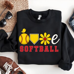 Peut inclure: Sweat-shirt noir avec un motif sur le thème du softball. Le motif comprend une balle de softball jaune avec un petit oiseau, un marbre jaune, une fleur jaune et le mot "SOFTBALL" en rouge. Le mot "love" est représenté par la balle de softball, le marbre, la fleur et la lettre "e".