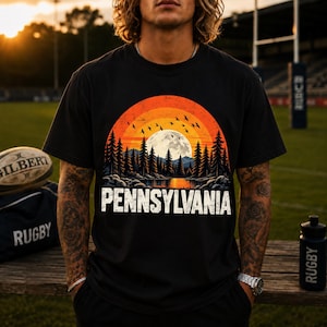 Peut inclure: Un t-shirt noir à manches courtes avec une impression graphique représentant un coucher de soleil sur une forêt et un lac, avec une pleine lune et des oiseaux. Le mot PENNSYLVANIA est imprimé en grandes lettres blanches.