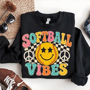 Peut inclure: Sweat-shirt noir avec un graphisme coloré qui dit "SOFTBALL VIBES". Le motif présente une balle de softball jaune avec des yeux en forme d'étoiles et un visage souriant, sur fond quadrillé, avec des signes de paix de chaque côté.