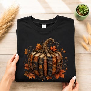 Peut inclure: Un sweat-shirt noir à col rond arborant un motif de citrouille composée de livres anciens aux dos ornés. La citrouille est entourée de feuilles d'automne détaillées et de vignes entrelacées, formant un design saisonnier sur le devant.