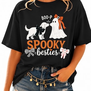 Peut inclure: Un t-shirt noir avec un motif représentant un chiot blanc avec un nœud noir et un fantôme blanc avec un nœud orange se touchant le nez. Le texte indique BOO-P et SPOOKY besties en orange et blanc, avec un nœud rayé noir et blanc et un nœud rose.