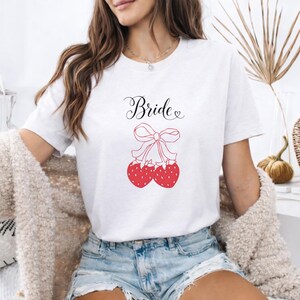 Könnte beinhalten: Weißes T-Shirt mit einem roten Erdbeermotiv und dem Wort "Bride" in Schreibschrift. Die Erdbeeren sind mit einer Schleife verbunden. Ein lässiges T-Shirt, geeignet für eine Hochzeitsveranstaltung.