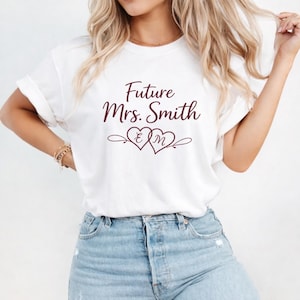 Może przedstawiać: Biała koszulka z podwiniętymi rękawami z napisem "Future Mrs. Smith" w kolorze bordowym. Poniżej dwa połączone serca z inicjałami "E" i "M". Koszulka jest połączona z niebieskimi dżinsami.